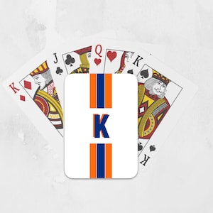 May include: A white playing card with a blue and orange stripe design and the letter "K" in the center. The card is surrounded by four other playing cards, including a King of Diamonds, a Jack of Clubs, a Queen of Hearts, and a King of Spades.