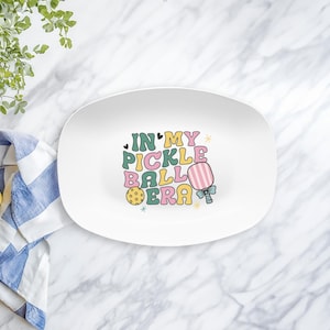 May include: White oval serving platter with the text "IN MY PICKLE BALL ERA" in colorful retro font. The design includes a pickleball paddle and ball. The platter is on a marble surface.