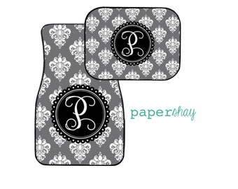 May include: A set of two gray car floor mats with a white damask pattern and a black circle with a white letter "P" in the center. The mats are for a car.