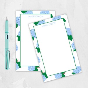 May include: Two white notepads with a blue hydrangea floral border and the name "JANIE DIXON" in light blue. A light teal fountain pen is positioned to the left of the notepads.