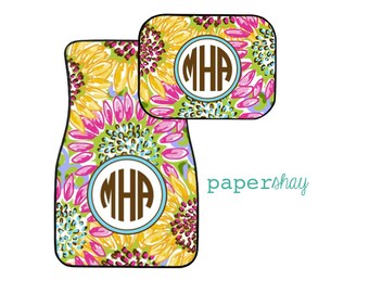 May include: Two car floor mats with a sunflower pattern in shades of yellow, pink, green, and purple. The mats have a monogram in a brown circle with the letters "MHA" on both mats. The text "papershay" is in turquoise.