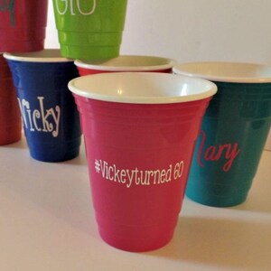 Op de afbeelding: Een verzameling kleurrijke plastic bekers, elk met een witte rand en gepersonaliseerde tekst. De bekers zijn in verschillende kleuren, waaronder rood, blauw, groen en roze. E&eacute;n beker heeft de tekst "#Vickeyturned60".