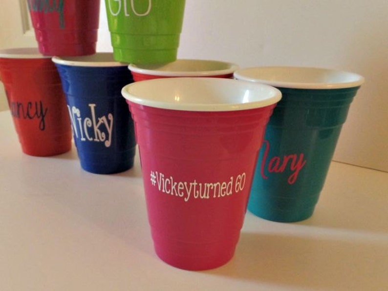 Op de afbeelding: Een verzameling kleurrijke plastic bekers, elk met een witte rand en gepersonaliseerde tekst. De bekers zijn in verschillende kleuren, waaronder rood, blauw, groen en roze. E&eacute;n beker heeft de tekst "#Vickeyturned60".