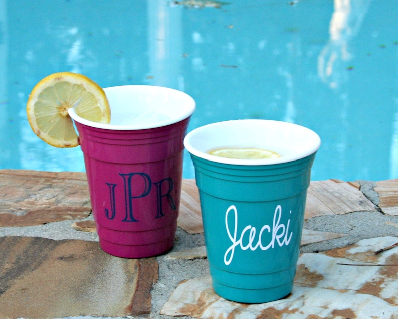 Op de afbeelding: Twee plastic bekers, &eacute;&eacute;n magenta en &eacute;&eacute;n turquoise, staan op een stenen oppervlak bij een zwembad. De magenta beker heeft de initialen "JPR" in marineblauw. De turquoise beker heeft de naam "Jacki" in witte letters. Beide bekers hebben een citroenschijfje.