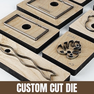 Könnte beinhalten: Eine Sammlung von Stanzformen mit verschiedenen Formen, darunter Rechtecke, Kurven und ein Monstera-Blatt-Design. Die Stanzformen sind aus Metall und auf Holzblöcken angeordnet. Der Text "CUSTOM CUT DIE" ist unten im Bild zu sehen.