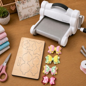May include: A white Big Shot die-cutting machine with a grey platform, a rectangular wooden cutting die with bow shapes, four small glittery fabric bows, a pair of pink-handled scissors, and a roll of light pink ribbon on a wooden surface.