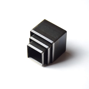 May include: Three nested square metal shapes in varying sizes. The shapes are dark grey with a silver accent around the edges. The objects are stacked together on a white background.