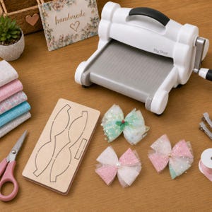 Op de afbeelding: Een witte Big Shot stansmachine, een houten stansvorm in de vorm van een strik, drie decoratieve haarstrikken van glinsterende tule, een schaar met roze handvatten en een stapel gevouwen stoffen vierkantjes op een houten ondergrond.