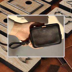 Könnte beinhalten: Eine kleine, rechteckige schwarze Ledertasche mit einer Handschlaufe. Die Tasche hat eine strukturierte Oberfläche und einen goldfarbenen Verschluss. Der Hintergrund umfasst Holzwerkzeuge und Lederstücke, was auf ein handgefertigtes Produkt hindeutet.