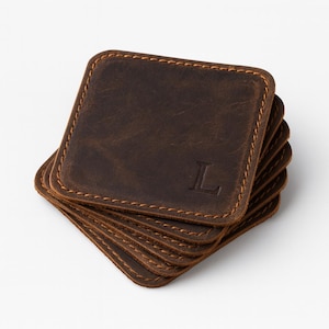 May include: A stack of square, brown leather coasters with stitched edges. Each coaster features the letter "L" embossed in the corner. The coasters are made of distressed leather, giving them a rustic appearance.