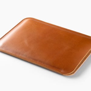 Peut inclure: Une pochette rectangulaire en cuir marron clair avec des coins arrondis. La pochette a une bordure cousue et une surface lisse et brillante. Conçu pour contenir une tablette ou un appareil similaire. Le cuir a un grain naturel.