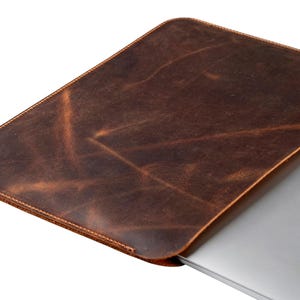 Könnte beinhalten: Eine braune Laptop-Hülle aus Leder mit einem Used-Look. Die Hülle ist rechteckig mit abgerundeten Ecken und einem genähten Rand. Ein silberner Laptop mit dem Apple-Logo ist teilweise sichtbar.
