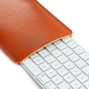 May include: A light brown leather keyboard sleeve partially covering a white keyboard. The sleeve is made of smooth leather and has a stitched edge. The keyboard has white keys with black lettering. The word "command" is visible on the keyboard.