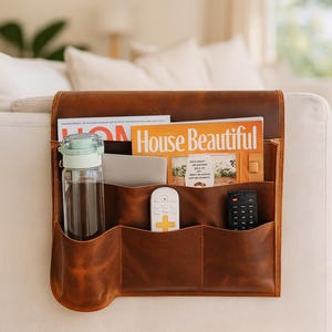 Könnte beinhalten: Ein brauner Leder-Couch-Organizer mit mehreren Taschen, der eine Zeitschrift, eine Wasserflasche, eine Fernbedienung und ein Tablet enthält. Der Organizer ist an der Seite eines weißen Sofas befestigt.