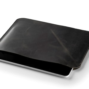 May include: A dark gray leather tablet sleeve with a tablet partially inserted. The sleeve has a smooth, slightly textured surface and rounded corners. The tablet has a silver frame and a black screen.