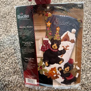 Op de afbeelding: Een Bucilla Black Bear Bonfire vilt kerstsok pakket in plastic verpakking. De set bevat bedrukt vilt, katoenen borduurgaren, pailletten, kralen en naalden om een kerstsok van 45,7 cm te maken met twee zwarte beren en een kampvuurscène.