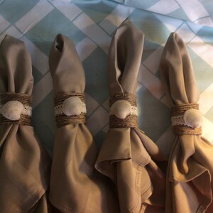May include: Four brown fabric napkins with burlap and seashell napkin rings. The rings are decorated with white lace.