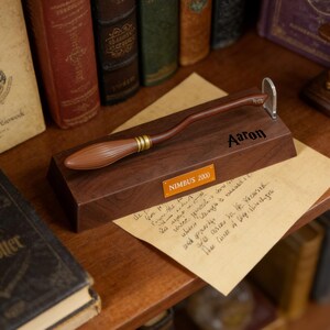 K&ouml;nnte beinhalten: Ein brauner Miniatur-Besen mit goldenen Akzenten, der auf einem dunklen Holzsockel ruht. Auf dem Sockel sind die Namen "Aaron" und "NIMBUS 2000" aufgedruckt. Unter dem Sockel befindet sich eine handschriftliche Notiz, im Hintergrund B&uuml;cher.