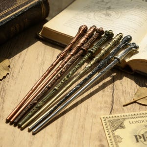 May include: Seven decorative wands in shades of copper, bronze, and silver, each with a unique design. The wands are displayed on a wooden surface, near an open book. The wands vary in length, with detailed patterns and textures.