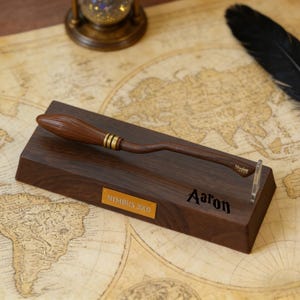 K&ouml;nnte beinhalten: Eine Holz-Besen-Replik, die auf einem dunklen Holzsockel ruht. Der Besen hat einen braunen Griff und Borsten mit goldfarbenen B&auml;ndern. Der Sockel hat eine goldene Plakette mit der Aufschrift "NIMBUS 2000" und der Name "Aaron" ist eingraviert.