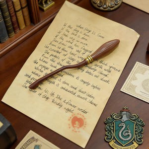 K&ouml;nnte beinhalten: Ein brauner und goldener dekorativer Besen-Requisit, der auf einem Vintage-Papier mit handschriftlichem Text liegt. Das Papier liegt auf einer Holzoberfl&auml;che, mit einem Hogwarts-Wappen und B&uuml;chern im Hintergrund.