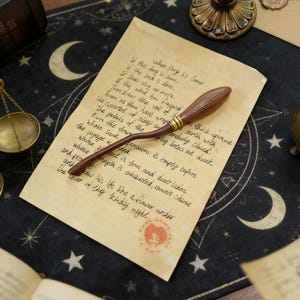 K&ouml;nnte beinhalten: Ein brauner Miniatur-Besen mit goldenen Akzenten liegt auf einem alten Papier mit handschriftlichem Text. Das Papier hat ein rotes Siegel und liegt vor einem dunklen Hintergrund mit Mond- und Sternmotiven.