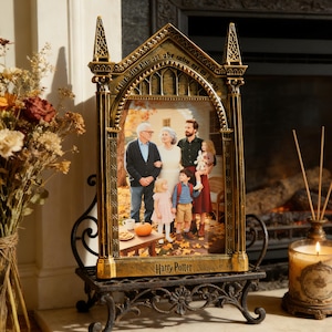 May include: A gold-colored Harry Potter picture frame with a family photo inside. The frame has an arched top with decorative detailing and sits on a black metal stand. A lit candle and dried flowers are in the background.