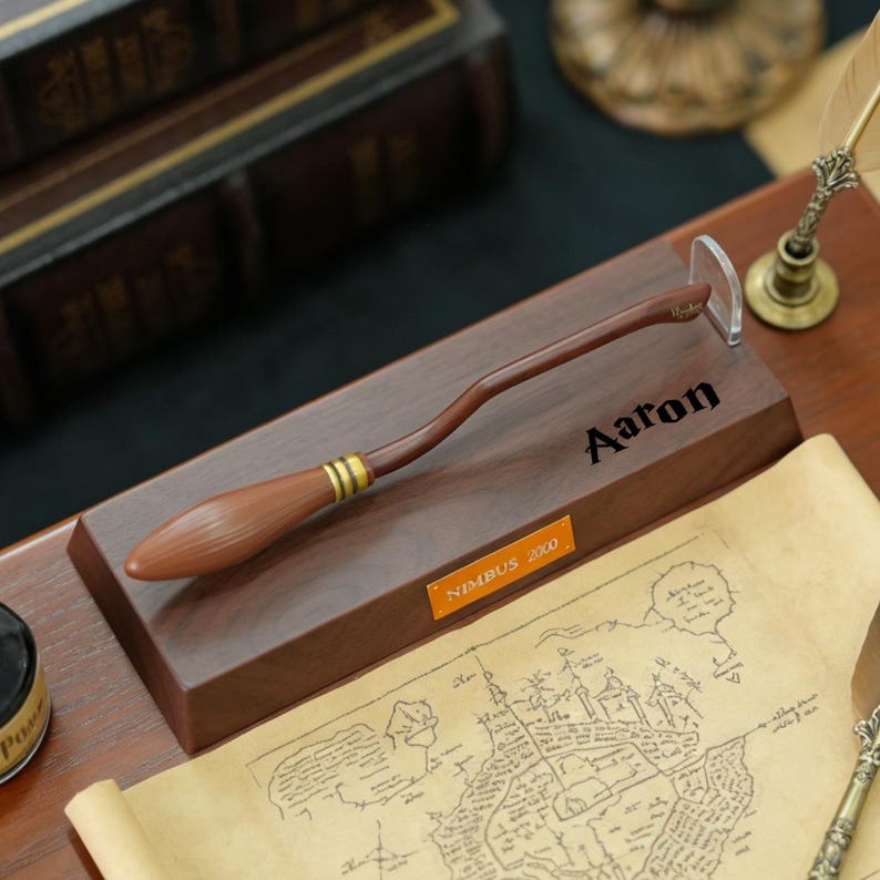 K&ouml;nnte beinhalten: Ein brauner Holzbesen mit goldenen Akzenten, der auf einem Holzsockel ruht. Der Sockel ist mit dem Namen "Aaron" und einem Schild mit der Aufschrift "Nimbus 2000" graviert. Eine Karte und eine Feder sind ebenfalls zu sehen.