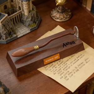 K&ouml;nnte beinhalten: Braune Nimbus 2000 Besenreplik mit goldenen Akzenten, pr&auml;sentiert auf einem Holzsockel. Der Sockel ist mit einem Namen und einer Plakette mit der Aufschrift "NIMBUS 2000" graviert. Ein klarer St&auml;nder st&uuml;tzt den Besen. Eine Miniatur-Burg ist im Hintergrund.