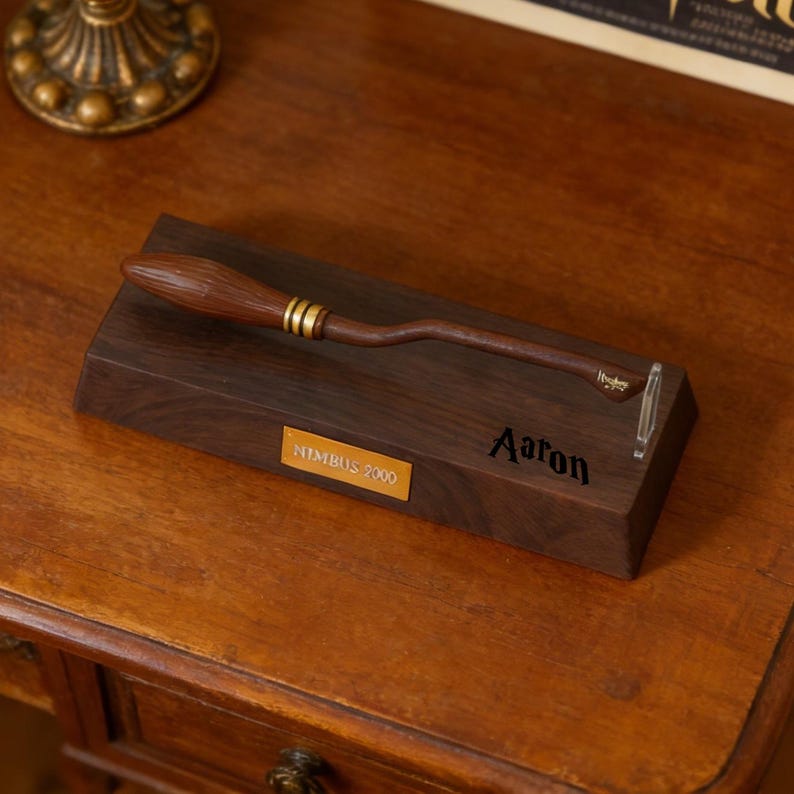 K&ouml;nnte beinhalten: Ein h&ouml;lzerner Nimbus 2000 Besen-Nachbau mit goldenen Akzenten, pr&auml;sentiert auf einem dunklen Holzsockel. Der Sockel hat ein goldenes Namensschild mit "Nimbus 2000" und dem Namen "Aaron". Ein klarer St&auml;nder st&uuml;tzt den Besen.