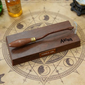 K&ouml;nnte beinhalten: Brauner Holz-Nimbus 2000 Besen-Nachbildung mit goldenen Akzenten, pr&auml;sentiert auf einem dunklen Holzsockel. Der Sockel ist mit dem Namen "Aaron" und einer Plakette mit der Aufschrift "Nimbus 2000" graviert. Der Hintergrund zeigt eine Himmelskarte.