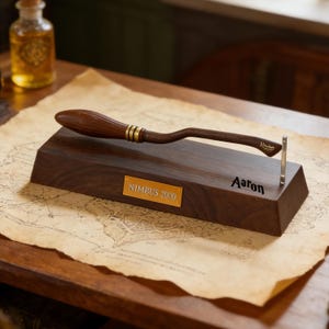 K&ouml;nnte beinhalten: Eine dunkelbraune Holz-Pr&auml;sentationsbasis mit einem Miniatur-Besen, der goldene Details aufweist. Die Basis hat eine goldene Plakette mit "NIMBUS 2000" und dem Namen "Aaron". Im Hintergrund ist eine kleine Flasche zu sehen.