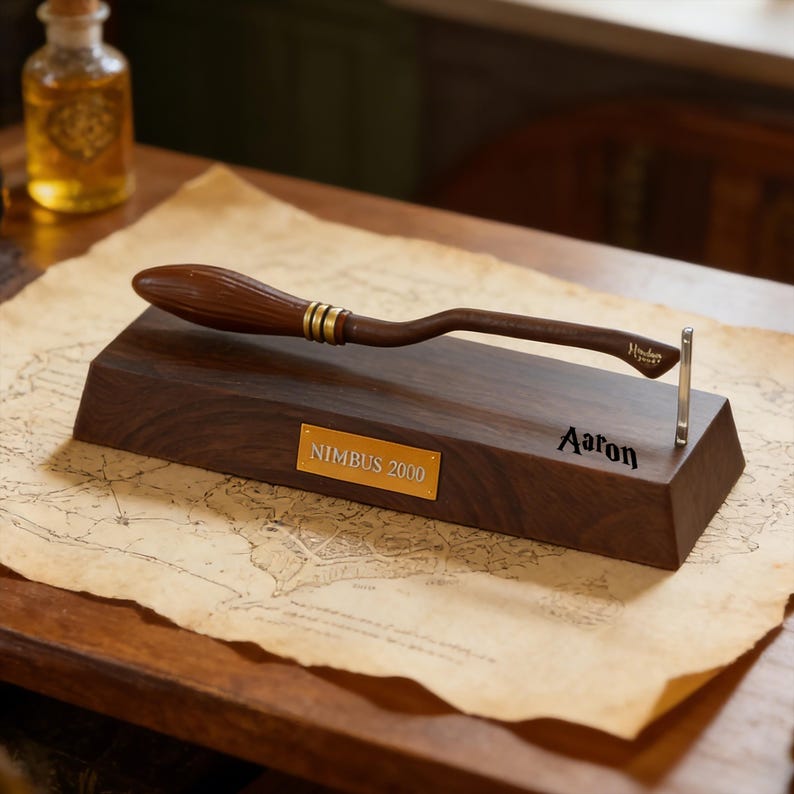 K&ouml;nnte beinhalten: Eine dunkelbraune Holz-Pr&auml;sentationsbasis mit einem Miniatur-Besen, der goldene Details aufweist. Die Basis hat eine goldene Plakette mit "NIMBUS 2000" und dem Namen "Aaron". Im Hintergrund ist eine kleine Flasche zu sehen.