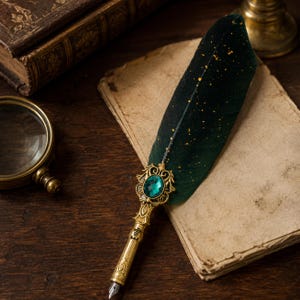 May include: A quill pen featuring a dark green feather with gold speckles, a gold-toned metal handle with ornate filigree detailing, and a central emerald-coloured oval gemstone. The handle is engraved with the name Emily and has a metal nib.