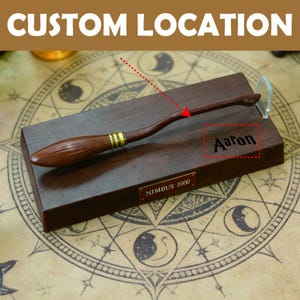 K&ouml;nnte beinhalten: Ein h&ouml;lzerner Besen mit goldenen Akzenten, der auf einem dunkelbraunen Sockel ruht. Der Sockel tr&auml;gt den eingravierten Namen "Aaron" und eine "Nimbus 2000"-Plakette. Oben steht "Custom Location".