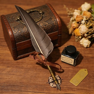 Puede incluir: Una pluma de estilo vintage con una pluma marrón y una punta de color bronce, atada con una cinta marrón. La pluma descansa sobre una superficie de madera junto a un pequeño cofre de madera, un tintero y una etiqueta de latón. La escena evoca la escritura.