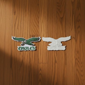 Op de afbeelding: Twee geborduurde stoffen patches met een groene adelaar met gespreide vleugels die een football vasthoudt. De linker patch toont het woord EAGLES in groene letters op een witte achtergrond, terwijl de rechter patch de effen witte achterkant toont.