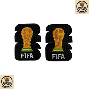 Op de afbeelding: Twee zwarte geborduurde patches met het cijfer 22 in een vetgedrukt lettertype. Gecentreerd op elke patch staat een goud en groen FIFA World Cup trofee-icoon. De witte tekst FIFA is onderaan elke patch met een zwarte rand geborduurd.