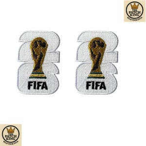 May include: Two embroidered patches featuring the FIFA World Cup trophy. The patches are white with a gold trophy design and the word "FIFA" in black. The patches are in the shape of the number "23".