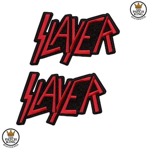 Puede incluir: Dos parches de Slayer. Cada parche presenta la palabra "SLAYER" en letras rojas estilizadas con un rayo a través de la "A", sobre un fondo negro. Los parches tienen un borde negro y están sobre un fondo blanco.