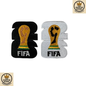 Puede incluir: Dos parches bordados con el número 2022, un gráfico de trofeo dorado y verde y el texto FIFA. Un parche tiene fondo negro con texto blanco, y el otro tiene fondo plateado con texto negro y detalles de trofeo en color dorado.