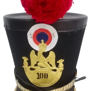 Może przedstawiać: Czarny kapelusz wojskowy z czerwonym pomponem. Posiada złoty emblemat z numerem 100, złotą opaskę i biało-czerwono-niebieski okrągły emblemat. Na rondzie widnieje napis "Hndmdprdetsindi".