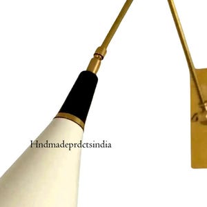 Könnte beinhalten: Eine wandmontierte, verstellbare Lampe mit einem konisch geformten, cremefarbenen Schirm. Die Lampe hat einen schwarzen und goldenen Akzent, einen Messingarm und eine rechteckige Wandhalterung. Das Design ist modern und minimalistisch.