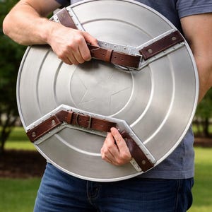 Op de afbeelding: Een zilverkleurig, rond schild met een stersymbool in het midden. Het schild heeft bruine leren riemen en metalen accenten. Het schild wordt door een persoon vastgehouden.