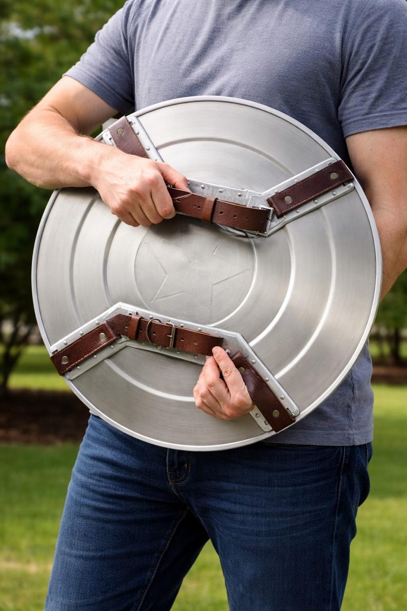 Op de afbeelding: Een zilverkleurig, rond schild met een stersymbool in het midden. Het schild heeft bruine leren riemen en metalen accenten. Het schild wordt door een persoon vastgehouden.