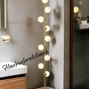 Puede incluir: Una lámpara de pie moderna con una estructura de metal dorado y múltiples bombillas esféricas de vidrio esmerilado. La lámpara está situada en un baño, junto a un lavabo y un espejo. El diseño es minimalista y elegante.