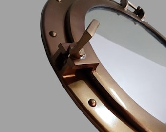 Könnte beinhalten: Ein bronzefarbenes, ovales Bullauge mit einer klaren Glasscheibe. Das Bullauge hat einen Klappverschluss und ist mit kleinen Nieten befestigt. Das Metall hat ein leicht verwittertes Aussehen, was auf ein nautisches oder industrielles Thema hindeutet.