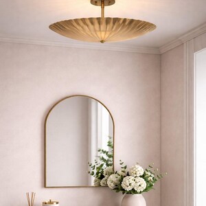 May include: A gold-toned, scalloped ceiling light fixture is mounted on a white ceiling. Below, a gold-framed arched mirror reflects a vase of white flowers and greenery. The room has a soft, neutral color palette.
