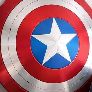 May include: A red, white, and blue shield with a large white star in the center. The shield has a circular design with concentric rings of red, silver, and blue. The shield is a replica of Captain America's shield.