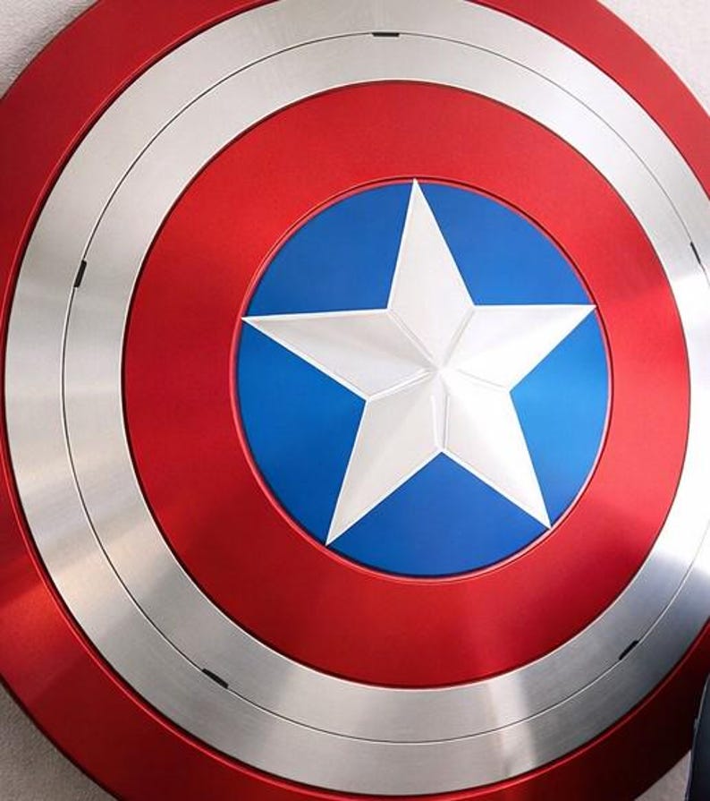 May include: A red, white, and blue shield with a large white star in the center. The shield has a circular design with concentric rings of red, silver, and blue. The shield is a replica of Captain America's shield.